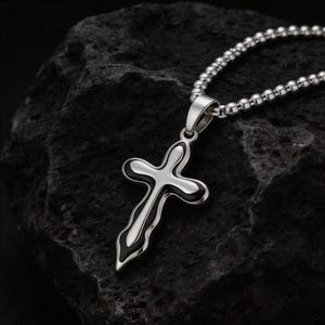 Peut inclure: Un pendentif en forme de croix argentée avec un contour noir est suspendu à une chaîne perlée. La croix a une forme allongée et stylisée. Le bijou est présenté sur un fond sombre et texturé.