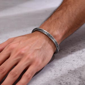 Pode incluir: Uma pulseira prateada com um design trançado. A pulseira é usada em um pulso, mostrando o design detalhado. O design da pulseira apresenta uma série de linhas angulares, criando uma aparência texturizada. Circunferência de aproximadamente 20 cm.