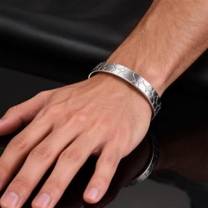 Pode incluir: Uma pulseira de punho prateada com um padrão orgânico texturizado. A pulseira é usada no pulso, mostrando seu design curvo e superfície reflexiva. A largura da pulseira é de aproximadamente 2 cm.