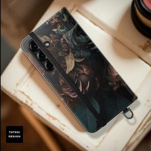 May include: A black phone case with a floral design featuring dark roses and leaves. The case has a black elastic band and a metal loop. The text "TOTESI DESIGN" is visible in the lower left corner.