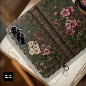 May include: A phone case with a vintage book design. The case features a dark green background with floral illustrations in shades of pink and white. The spine of the book is visible, along with the text "TOTESI DESIGN".