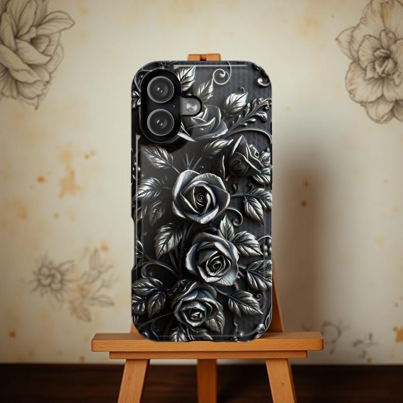 May include: Black phone case with a 3D rose design. The case features a detailed relief of roses and leaves in shades of gray and silver. The phone case is displayed on a small wooden easel.