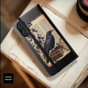 May include: A black leather-textured phone case featuring a rectangular insert with a vintage book page background, a black raven perched on a stack of books, and floral illustrations. A black elastic band secures the case, which has a camera cutout.