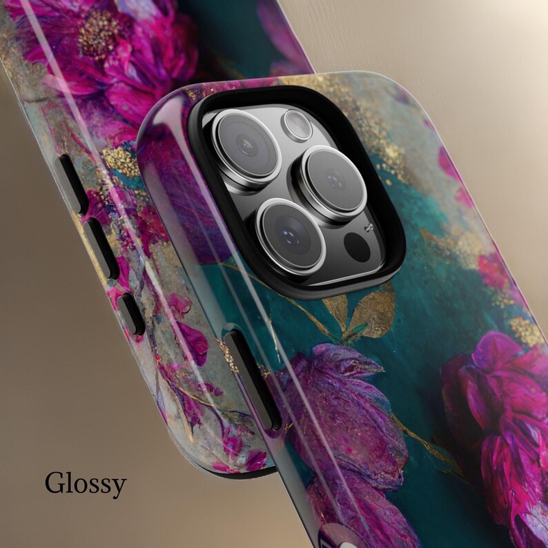 May include: A glossy phone case with a floral design featuring pink and purple flowers on a teal background with gold accents. The case has a raised camera bump with a black border.
