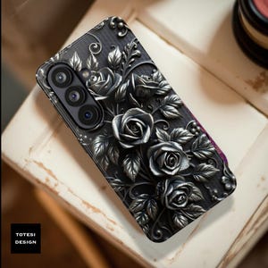 May include: Black phone case with a raised silver rose design. The case features a detailed floral pattern with leaves and roses. The phone case has a camera lens and the text "TOTESI DESIGN" in a black box.