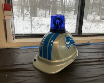 May include: A gray helmet with blue stripes and a blue Detroit Lions logo. A blue light is mounted on top of the helmet. The helmet is sitting on a black surface.
