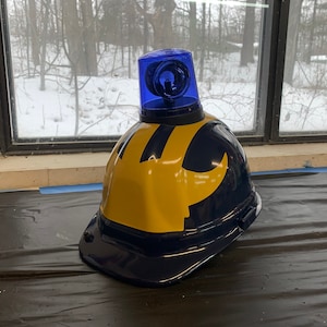 May include: A blue hard hat with a yellow 'M' logo and a blue flashing light on top.
