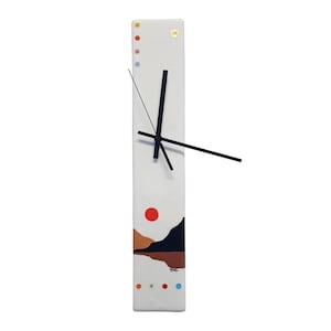 May include: A rectangular white wall clock with black hands. The clock face features a landscape design with a red sun, brown and black mountains, and colorful dots. The clock is approximately 61 cm long.