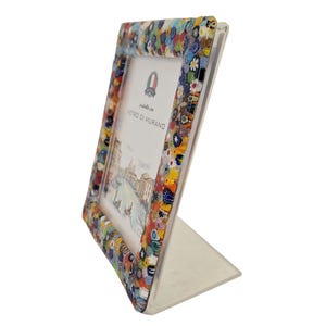 May include: A colorful, decorative picture frame with a mosaic-style border. The frame features a variety of small, multi-colored glass pieces. The frame is supported by a clear, angled stand and holds a picture with the text "Vetro di Murano".