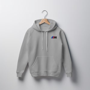 May include: A light grey hooded sweatshirt with a front pocket and white drawstrings. The sweatshirt features a small logo on the upper left chest with blue, red, and black colours.