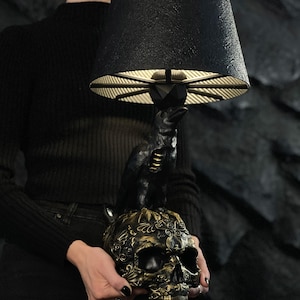Puede incluir: Lámpara de mesa negra con un cuervo posado sobre una base de calavera con detalles dorados. La lámpara tiene una pantalla negra y texturizada.