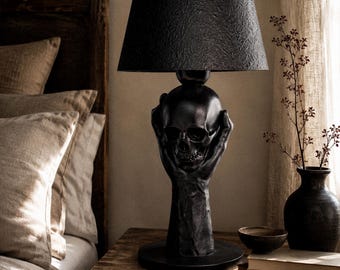 Peut inclure: Une lampe de table noire dont le pied représente deux mains tenant un crâne humain. La lampe est dotée d'un abat-jour tambour noir texturé et repose sur une base circulaire. Le design est monochrome avec une finition mate sur la sculpture.