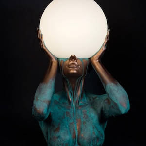 Puede incluir: Una l&aacute;mpara de mesa escultural que presenta un busto en tonos bronce con un acabado de p&aacute;tina turquesa desgastada. La figura tiene ambos brazos levantados, con las manos sosteniendo una gran esfera de vidrio blanco esmerilado.
