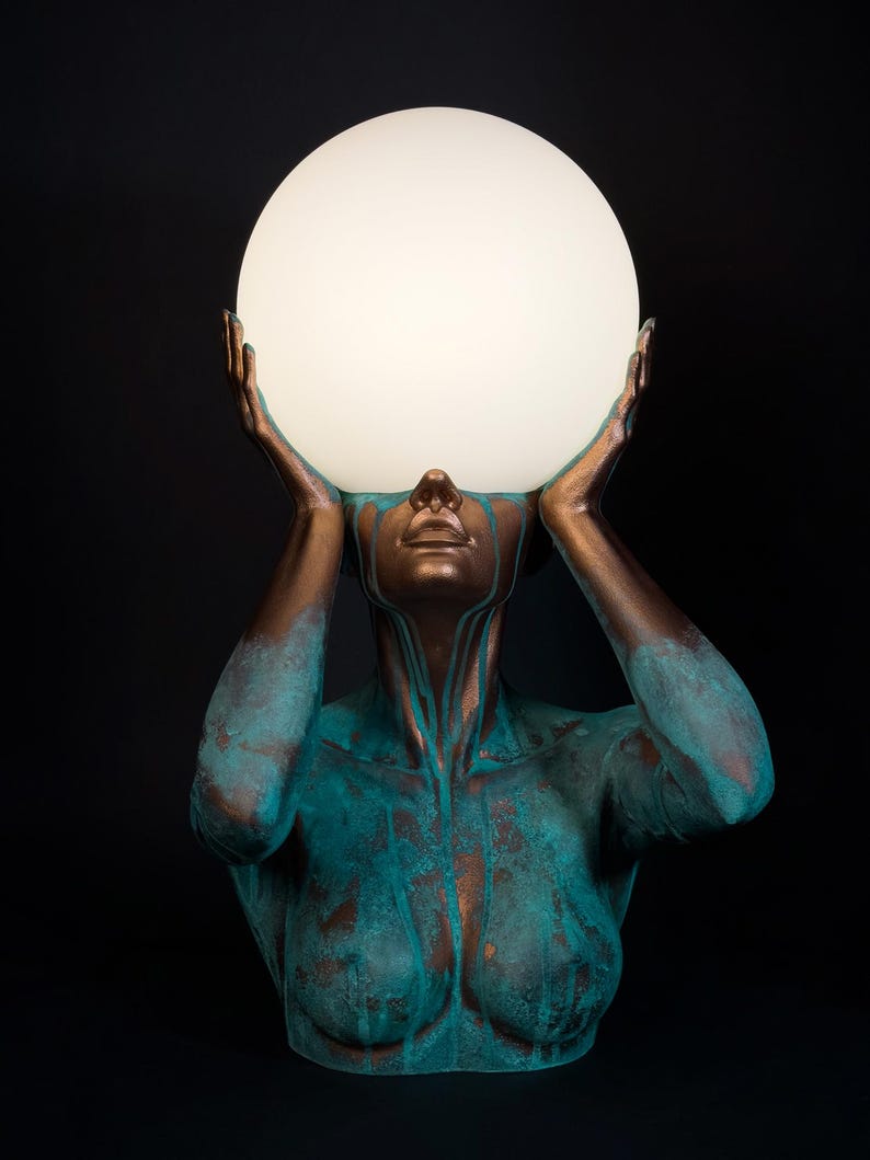 Puede incluir: Una l&aacute;mpara de mesa escultural que presenta un busto en tonos bronce con un acabado de p&aacute;tina turquesa desgastada. La figura tiene ambos brazos levantados, con las manos sosteniendo una gran esfera de vidrio blanco esmerilado.