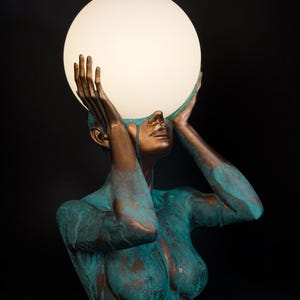 Puede incluir: Una l&aacute;mpara de mesa escultural que presenta un busto en tonos bronce con un acabado de p&aacute;tina verde azulada desgastada. La figura tiene ambos brazos levantados, con las manos sosteniendo una gran esfera de cristal blanco iluminada.