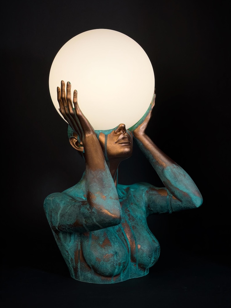 Puede incluir: Una l&aacute;mpara de mesa escultural que presenta un busto en tonos bronce con un acabado de p&aacute;tina verde azulada desgastada. La figura tiene ambos brazos levantados, con las manos sosteniendo una gran esfera de cristal blanco iluminada.