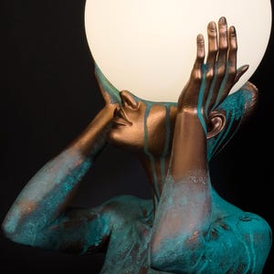 Puede incluir: Una escultura de una figura con acabado en bronce y gotas de p&aacute;tina color cerceta, sosteniendo una gran l&aacute;mpara esf&eacute;rica de cristal blanco iluminada sobre su cabeza. La figura se muestra desde el pecho, con las manos levantadas.