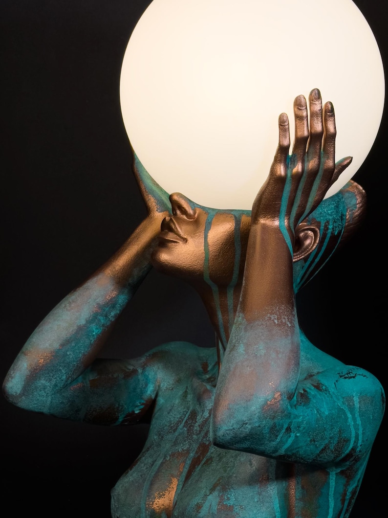 Puede incluir: Una escultura de una figura con acabado en bronce y gotas de p&aacute;tina color cerceta, sosteniendo una gran l&aacute;mpara esf&eacute;rica de cristal blanco iluminada sobre su cabeza. La figura se muestra desde el pecho, con las manos levantadas.
