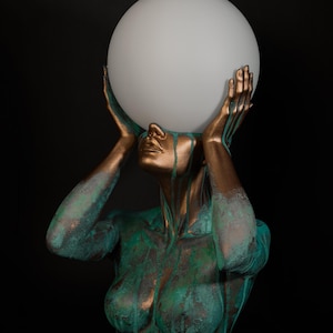 Puede incluir: Una l&aacute;mpara de mesa escultural que presenta un busto en tonos bronce con un acabado de p&aacute;tina verde desgastada. La figura tiene ambas manos levantadas para sostener un gran globo esf&eacute;rico de vidrio blanco esmerilado como luz.