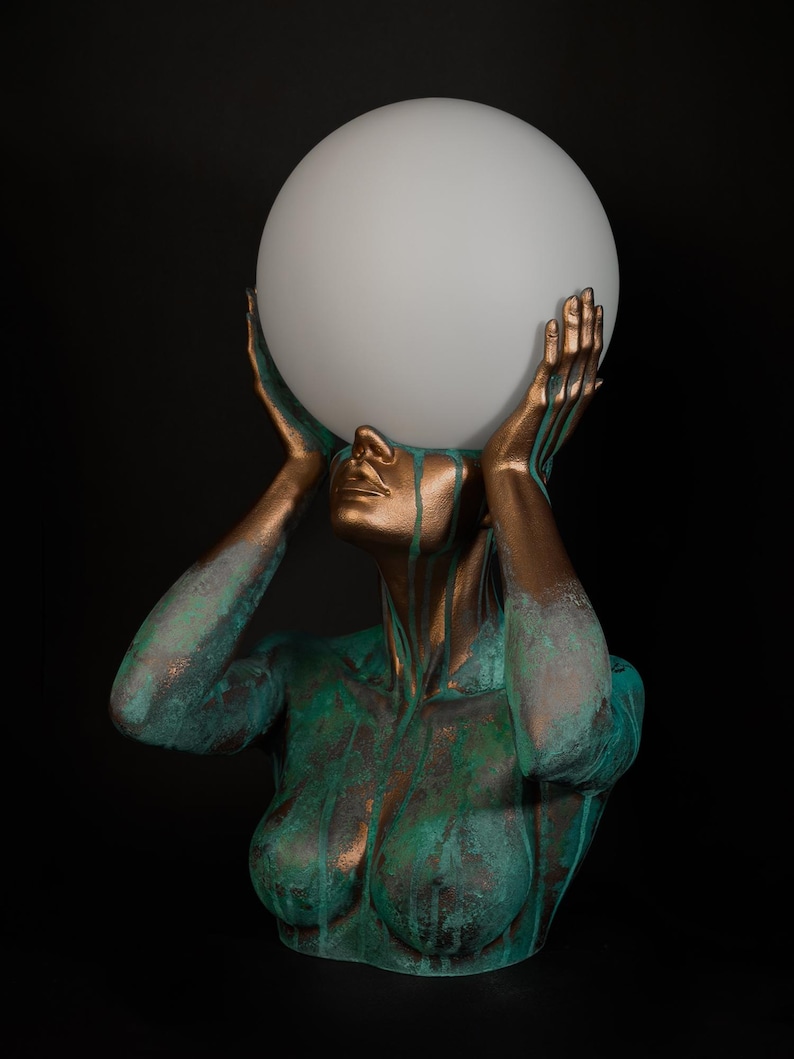 Puede incluir: Una l&aacute;mpara de mesa escultural que presenta un busto en tonos bronce con un acabado de p&aacute;tina verde desgastada. La figura tiene ambas manos levantadas para sostener un gran globo esf&eacute;rico de vidrio blanco esmerilado como luz.