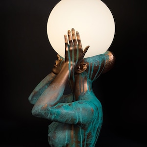 Puede incluir: Una l&aacute;mpara de mesa escultural que presenta un busto en tonos bronce con un acabado de p&aacute;tina turquesa. La figura tiene las manos levantadas hacia el rostro, sosteniendo un gran globo de luz esf&eacute;rico de vidrio blanco esmerilado.