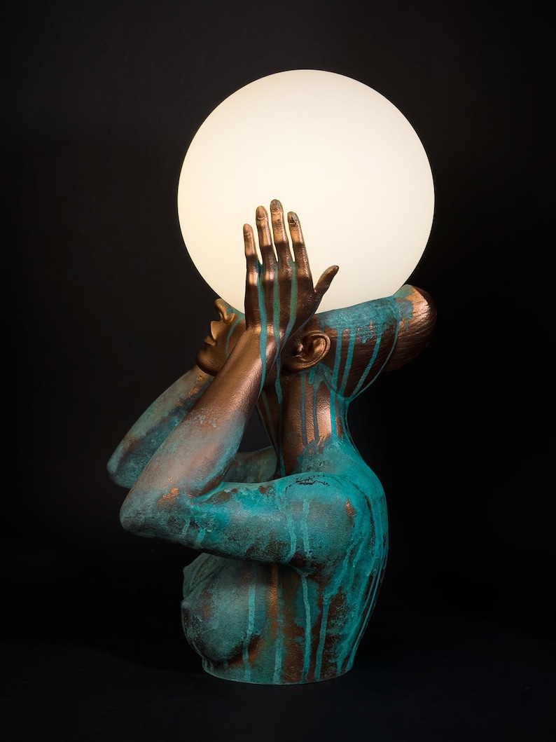 Puede incluir: Una l&aacute;mpara de mesa escultural que presenta un busto en tonos bronce con un acabado de p&aacute;tina turquesa. La figura tiene las manos levantadas hacia el rostro, sosteniendo un gran globo de luz esf&eacute;rico de vidrio blanco esmerilado.