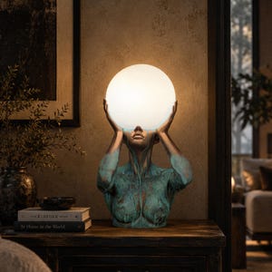 Puede incluir: Una l&aacute;mpara de mesa escultural que presenta un busto en tonos bronce con un acabado de p&aacute;tina verde desgastada. La figura sostiene una gran esfera de cristal blanco brillante sobre su cabeza, que sirve como fuente de luz para la pieza.