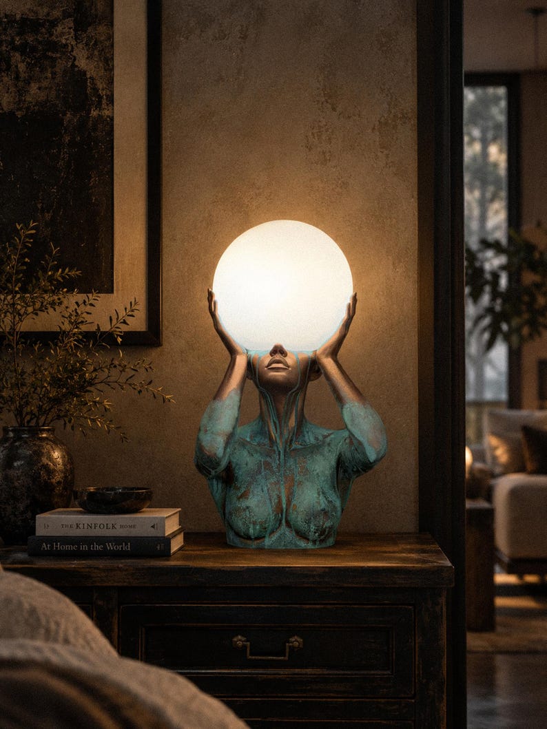 Puede incluir: Una l&aacute;mpara de mesa escultural que presenta un busto en tonos bronce con un acabado de p&aacute;tina verde desgastada. La figura sostiene una gran esfera de cristal blanco brillante sobre su cabeza, que sirve como fuente de luz para la pieza.