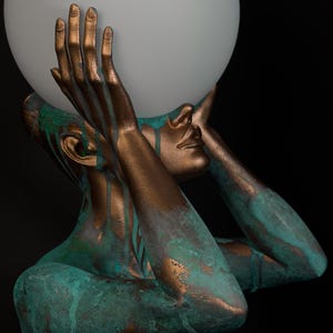 Puede incluir: Una l&aacute;mpara de mesa escultural que presenta una figura en tonos bronce con un acabado de p&aacute;tina verde azulado desgastado. La figura sostiene una gran esfera de vidrio blanco esmerilado sobre su cabeza con ambas manos, creando luz.