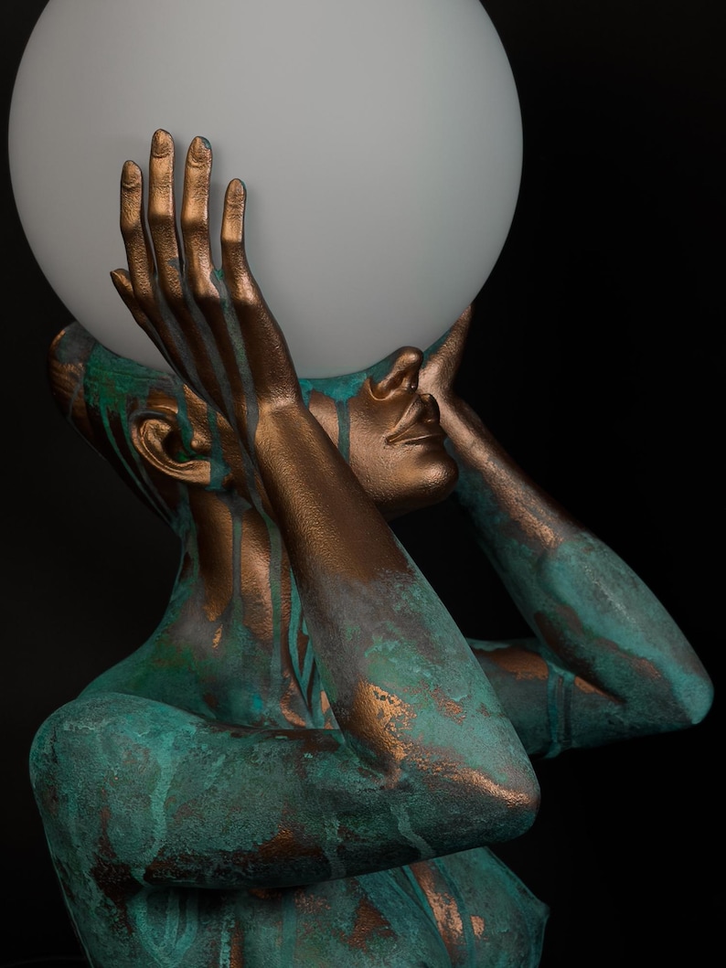 Puede incluir: Una l&aacute;mpara de mesa escultural que presenta una figura en tonos bronce con un acabado de p&aacute;tina verde azulado desgastado. La figura sostiene una gran esfera de vidrio blanco esmerilado sobre su cabeza con ambas manos, creando luz.