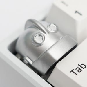 May include: A close-up shot of a white mechanical keyboard featuring a silver robot-shaped keycap on the 'Tab' key. The keycap has two circular eye-like features. The keyboard keys are white with black lettering.