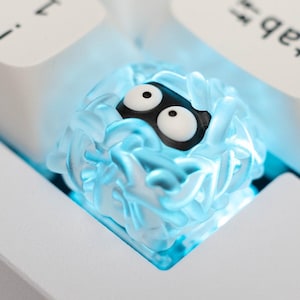 May include: A clear blue keycap with a white and black cartoon face. The keycap is glowing blue and is on a white keyboard.