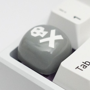 Op de afbeelding: Close-up van een wit toetsenbord met een grijze keycap met een schedel en gekruiste botten symbool en een witte "X". De "Tab"-toets is zichtbaar. Het toetsenbord staat tegen een witte achtergrond.