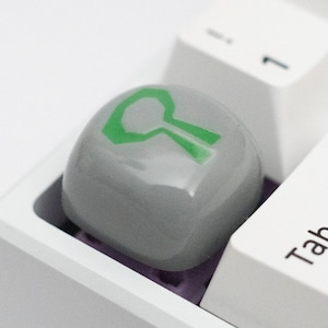 Op de afbeelding: Een wit toetsenbord met een grijze toets waarop een groen symbool is gedrukt. De toets is gelabeld met "Tab".
