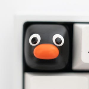 May include: A black penguin-shaped keycap with white eyes and an orange beak is installed on a white keyboard. The keyboard also has white keys with the numbers 1 and 2, and the Tab and Q keys.