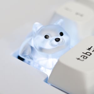 May include: Close-up of a white mechanical keyboard with a custom keycap shaped like a clear, translucent polar bear. The bear keycap is lit up, and the keyboard keys are white with black lettering.