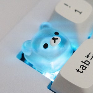 May include: A white keyboard with a blue light shining from underneath the keys. The 'tab' key is highlighted and a blue, clear, bear-shaped keycap is on the key to the left of the 'tab' key.