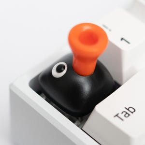 May include: A black and orange keycap shaped like a cartoon character, with a white eye, sits on a white keyboard. The keycap replaces the Tab key. The keyboard has white keys and a white frame.