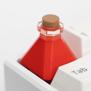Puede incluir: Una tecla roja en forma de poción con un tapón marrón se encuentra en un teclado blanco. La tecla tiene un diseño geométrico y está colocada en la tecla "Tab". El teclado es blanco.