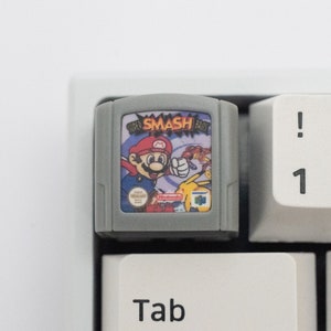Könnte beinhalten: Eine graue Nintendo 64-Spielkassette mit einem bunten Bild von Mario und Pikachu auf der Vorderseite. Die Kassette ist mit "Super Smash Bros." beschriftet.