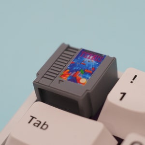 Puede incluir: Un teclado gris y blanco con una tecla personalizada que presenta un diseño de cartucho de videojuego retro. La tecla tiene una etiqueta que dice "TETRIS".