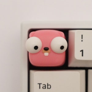 Könnte beinhalten: Eine rosafarbene Tastaturkappe mit einem Comic-Gesicht und großen weißen Augen. Die Kappe befindet sich auf einer weißen Tastatur mit Tasten, die mit "Tab", "Q", "1", "!", "2" und "@" beschriftet sind.