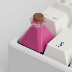 Puede incluir: Una tecla magenta en forma de pirámide, que se asemeja a una botella de poción, con una tapa marrón, descansa sobre un teclado blanco. La tecla está colocada entre las teclas "Tab" y "Caps Lock".