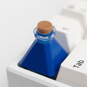 Puede incluir: Una botella azul en forma de pirámide con un corcho marrón se encuentra sobre un teclado blanco. La botella es de color azul vibrante y tiene una parte superior transparente facetada. La tecla del teclado al lado de la botella dice "Tab".