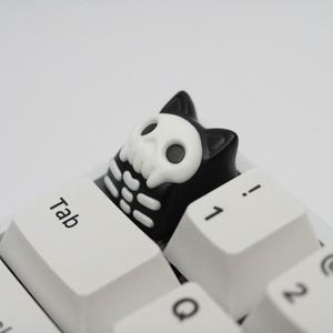 May include: A black and white keyboard keycap with a skull design. The keycap is shaped like a cartoon character and has a white skull with black eyes and a black outline. The keycap is sitting on a white keyboard.