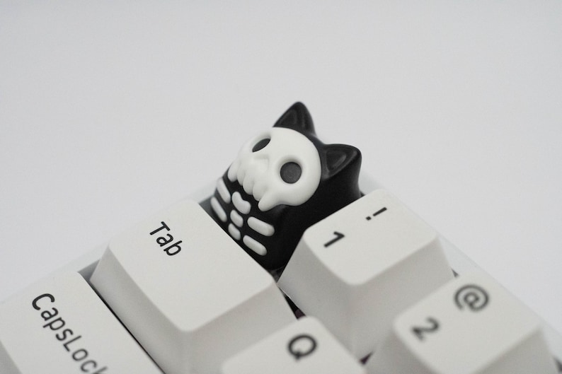May include: A black and white keyboard keycap with a skull design. The keycap is shaped like a cartoon character and has a white skull with black eyes and a black outline. The keycap is sitting on a white keyboard.