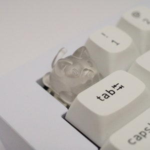 May include: A white keyboard with a clear plastic cat keycap on the tab key. The keycap is shaped like a cat with a cute face.