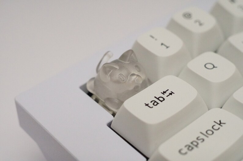 May include: A white keyboard with a clear plastic cat keycap on the tab key. The keycap is shaped like a cat with a cute face.