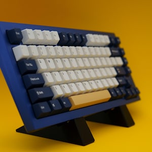 May include: A blue and black mechanical keyboard with white keycaps and a gold space bar. The keyboard is on a black stand.