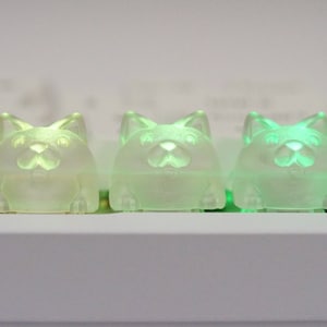 May include: Three translucent green cat-shaped keycaps with a light green glow. The keycaps are arranged in a row on a white surface.